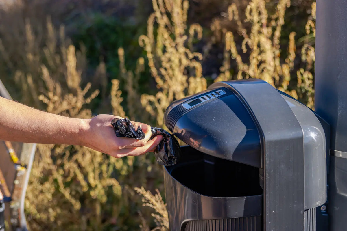 Hand disposing sustainable dog waste bags in outdoor trash bin near vegetation