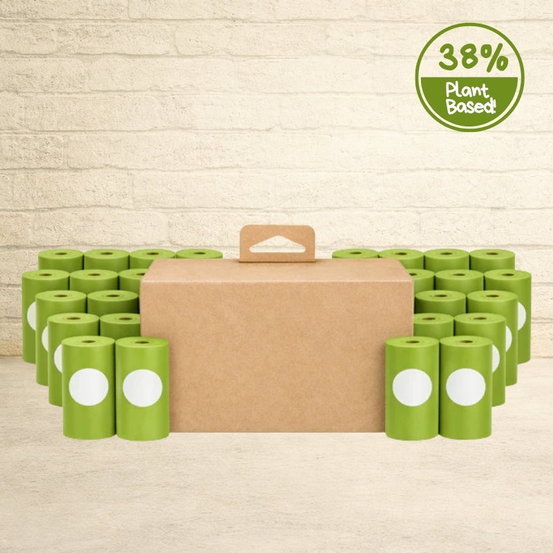 Brown cardboard case of Original Poop Bags surrounded by green 38% plant-based dog waste bag rolls on a wooden background.