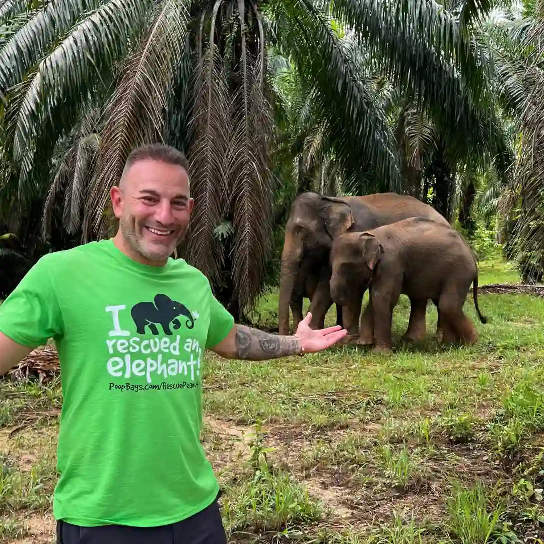 Man in green elephant rescue shirt posing with rescued elephants in tropical sanctuary setting